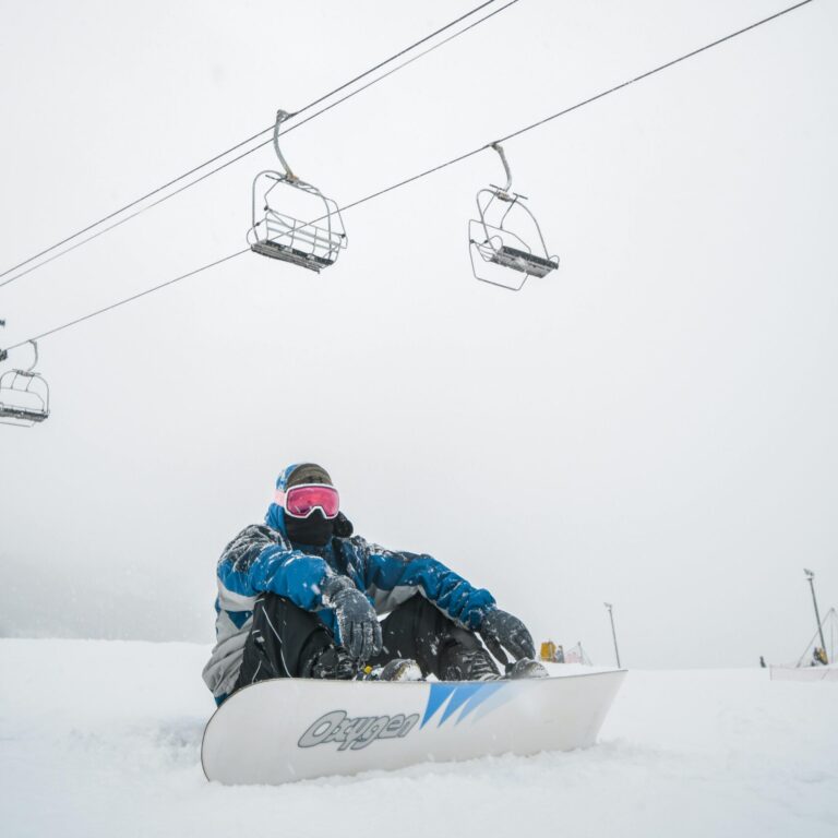 Snowboarder in winter gear resting on a snowy slope under cable cars in a ski resort.